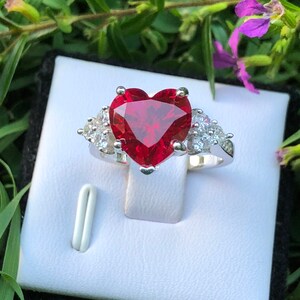 May include: A silver ring with a large red heart-shaped gemstone surrounded by smaller clear gemstones.