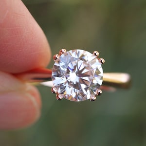 May include: A close-up of a rose gold engagement ring with a large, round, clear diamond.
