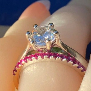 May include: A silver ring with a large, clear, round diamond set in a six-prong setting. The ring has a thin band with a row of small pink gemstones.