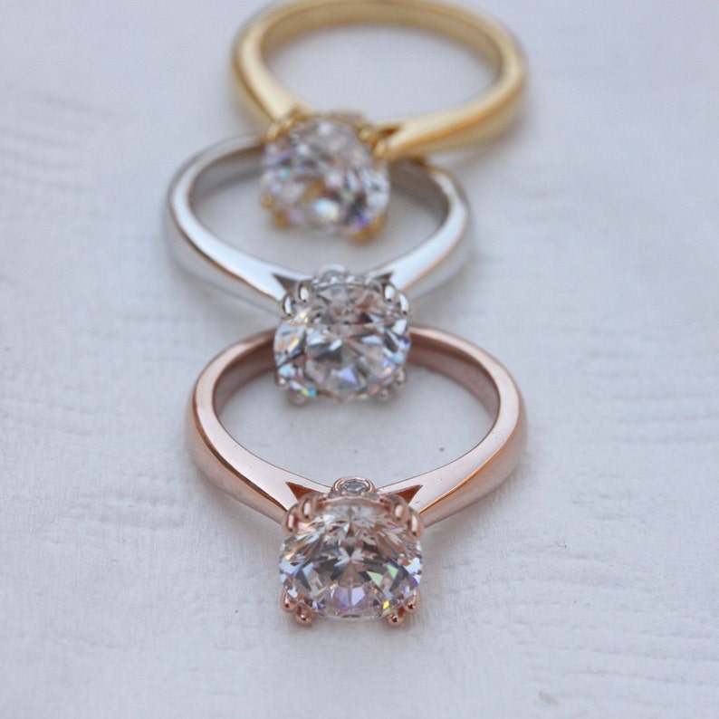 May include: Three engagement rings with different metal settings: gold, silver, and rose gold. Each ring features a round, clear diamond.