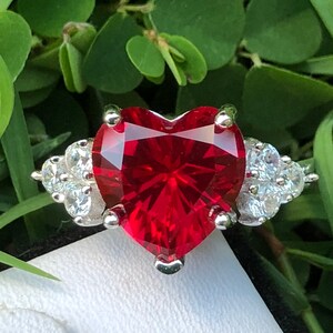 May include: A silver ring with a large, heart-shaped, red gemstone surrounded by smaller, clear gemstones.