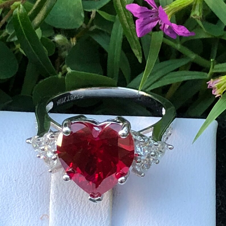 May include: A silver ring with a large heart-shaped red gemstone surrounded by smaller clear gemstones. The ring is marked with the text "MU215255".