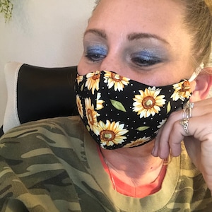 May include: A woman wearing a black face mask with a sunflower pattern. The mask has white polka dots and is worn over her nose and mouth. She is wearing a camouflage shirt and has blue eyeshadow on.