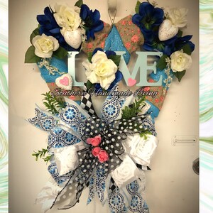 Könnte beinhalten: Ein herzförmiger Kranz mit einem blau-weiß karierten Band, weißen Rosen, blauen Blumen und dem Wort "LOVE" in blauen Buchstaben. Der Kranz ist mit weißen und rosa Herzen verziert. Der Text "Southern Handmade Living" ist am unteren Rand des Kranzes sichtbar.