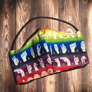 May include: A rainbow-colored face mask with the letters of the alphabet in American Sign Language printed on it. The mask has a black elastic band.