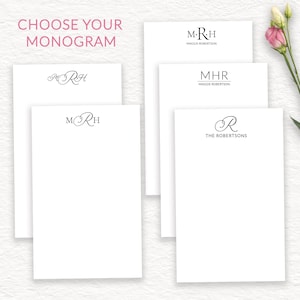 May include: White stationery with various monogram designs. The text "CHOOSE YOUR MONOGRAM" is at the top left. The stationery features initials and names in elegant fonts. A pink and white flower is in the upper right corner.