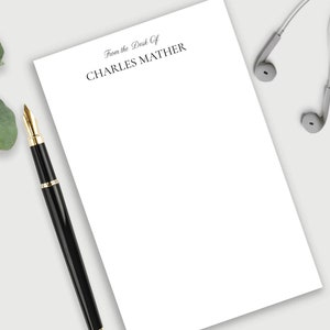 May include: A white notepad with the text "From the Desk of CHARLES MATHER" printed at the top. A black pen and white earbuds are on a white surface next to the notepad.