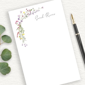 May include: A white notepad with a floral design in shades of purple, pink, yellow, and green. The name "Sarah Rivers" is written in cursive at the top of the notepad.