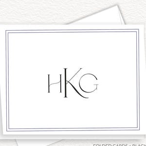 May include: White folded cards with a black border and the initials "HKG" in a serif font. The cards are stacked on top of each other. A black and gold pen is on the right side of the cards. The text "FOLDED CARDS • BLACK INK" is at the bottom.
