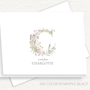 May include: White stationery set featuring a floral letter "C" design. The letter is composed of watercolor flowers in shades of pink, purple, and yellow. The text "a note from CHARLOTTE" is printed below the floral design.