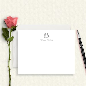 May include: A white stationery card featuring a black horseshoe icon centered above the printed name Melissa Mellon. The card is accompanied by a matching white envelope, a single pink rose with a green stem, and a black fountain pen.