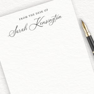 May include: A white notepad with the text "From the desk of Sarah Kensington" printed at the top. A black pen is lying on the notepad.