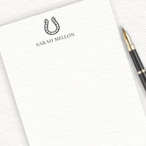 May include: A white notepad with a black horseshoe design and the text "SARAH MELLON" printed on the top. A black pen with gold accents is lying on the notepad.
