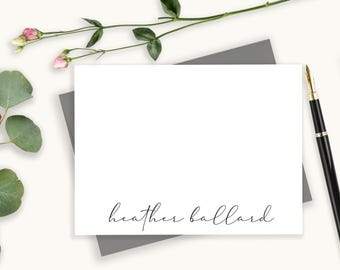 May include: A white rectangular stationery card featuring the name heather ballard printed in a black cursive script at the bottom, resting on a gray envelope next to a black fountain pen and a sprig of green leaves with pink flowers.