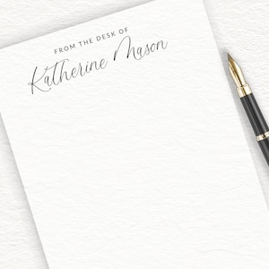 May include: A white sheet of stationery with the text FROM THE DESK OF Katherine Mason printed in black ink, accompanied by a black fountain pen with a gold nib, green eucalyptus leaves, and small pink flower buds.