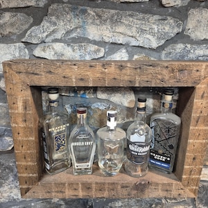 Puede incluir: Una estantería rústica de madera exhibe cinco botellas de licor vacías contra una pared de piedra. Las botellas varían en forma y tamaño, con etiquetas como "Heaven's Door" y "Angel's Envy". La estantería está hecha de madera reciclada.