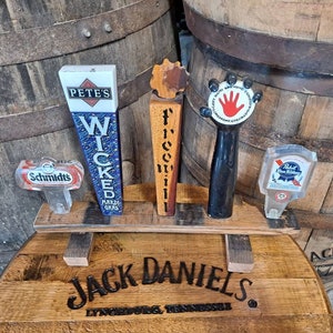May include: A collection of beer tap handles displayed on a wooden stand. The handles feature various designs and brand names, including "Pete's Wicked," "Freeville," and "Left Hand Brewing Company." The stand is placed on a Jack Daniel's barrel.