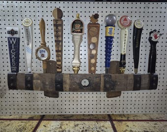 May include: A horizontal wooden barrel stave wall mount holding nine beer tap handles. The handles feature various brands including Pete's Wicked, Warsteiner, Miller Lite, Freewill, Smithwick's, and Bass, with diverse shapes, colors, and materials.