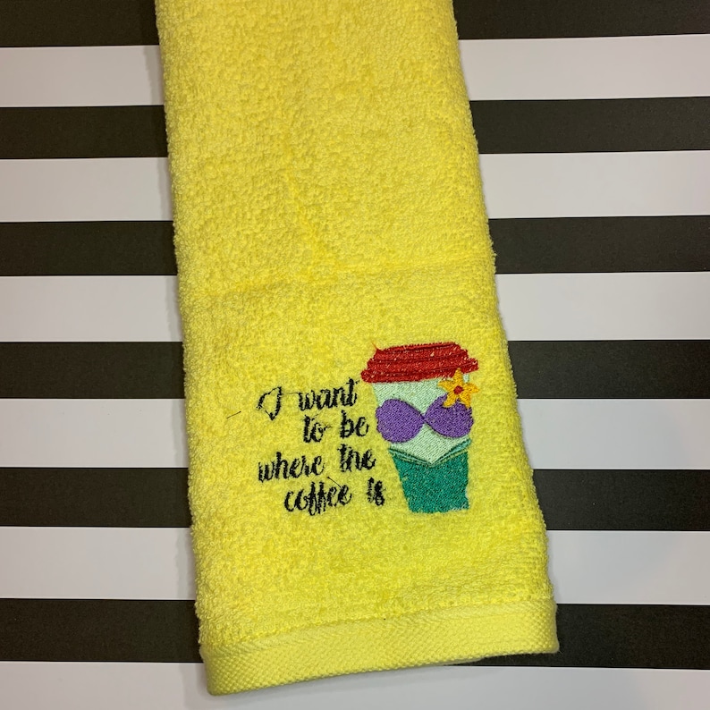 May include: A bright yellow hand towel featuring an embroidered design of a coffee cup wearing a purple seashell bra and a yellow flower. The text reads, I want to be where the coffee is, stitched in black thread next to the coffee cup graphic.
