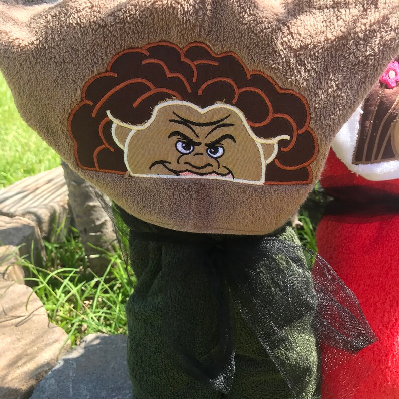 May include: A tan hooded towel featuring an embroidered character face with dark brown curly hair, thick eyebrows, and a smirk. The hood is attached to a dark green towel body, accented with a sheer black ribbon tied around the neck area.