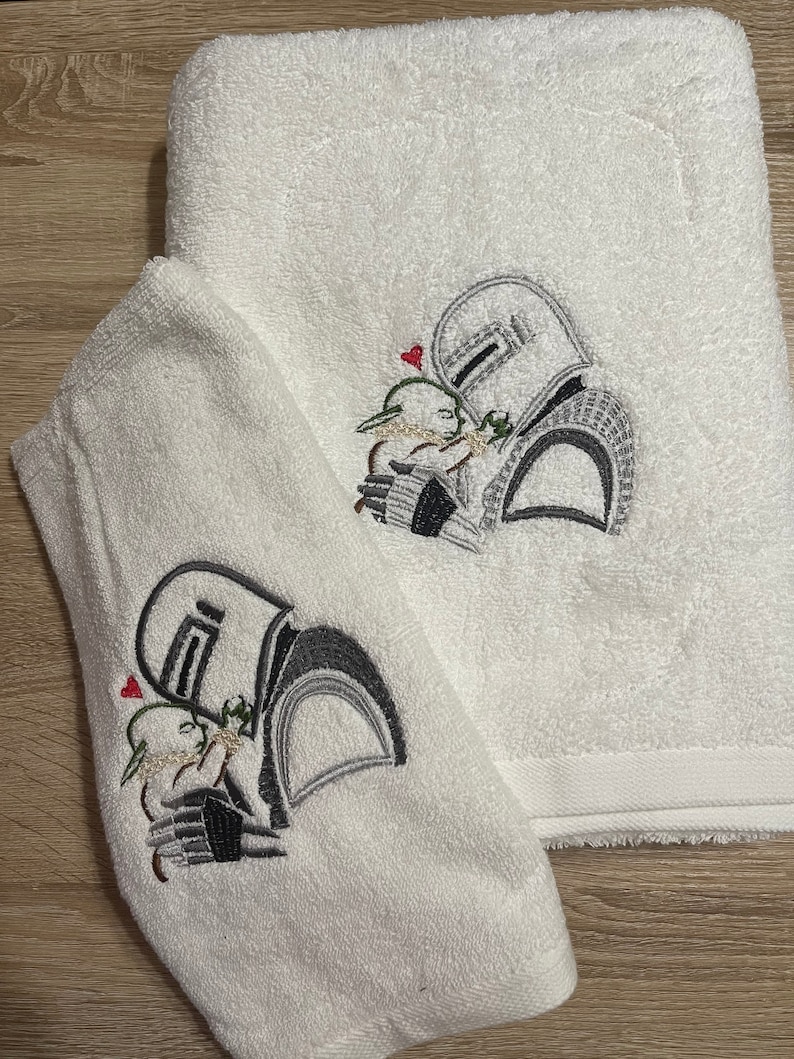 May include: Two white terry cloth towels featuring an embroidered design of a helmeted figure holding a small green creature. The embroidery uses shades of gray, black, and green thread, with a small red heart positioned above the figures.