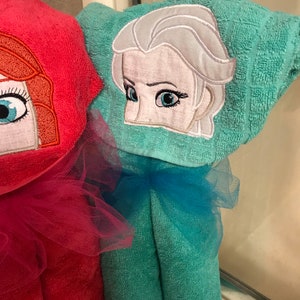 May include: A bright red towel and a teal towel, each featuring an embroidered character face on the hood. The teal towel displays a character with platinum blonde hair and blue eyes, while the red towel shows a character with orange hair.