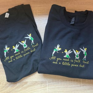 May include: Two black long-sleeved shirts featuring embroidered designs of four Tinker Bell figures in green and yellow. Below the figures, the text reads: All you need is faith, trust and a little pixie dust, stitched in yellow thread.