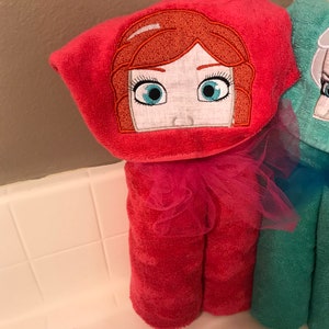 May include: A bright pink hooded towel featuring an embroidered face with red hair and blue eyes. The hood is accented with a pink tulle ruffle at the neck, and the towel is made of a soft, textured terry cloth fabric with a vibrant solid colour.