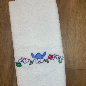 May include: A white rectangular terry cloth towel featuring an embroidered design near the bottom edge. The embroidery includes a blue character silhouette, a red surfboard, a red hibiscus flower, a green leaf, and brown accents on a blue scrollwork.