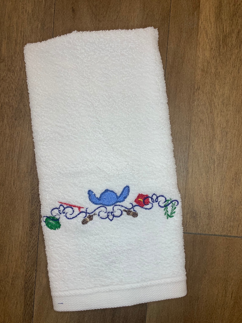 May include: A white rectangular terry cloth towel featuring an embroidered design near the bottom edge. The embroidery includes a blue character silhouette, a red surfboard, a red hibiscus flower, a green leaf, and brown accents on a blue scrollwork.
