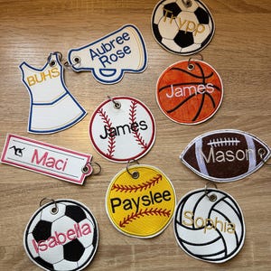 May include: A collection of personalized sports-themed keychains. Designs include a soccer ball, basketball, baseball, football, and a cheerleader megaphone. Each keychain features a name embroidered in a contrasting color.