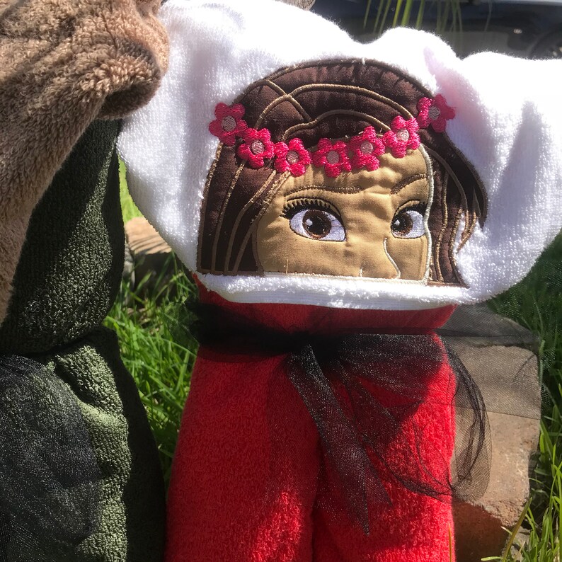 May include: A white hooded towel featuring an embroidered face of a character with brown hair and a pink flower crown. The towel has a red body section and is accented with a black tulle ribbon tied around the neck area, positioned next to a green towel.