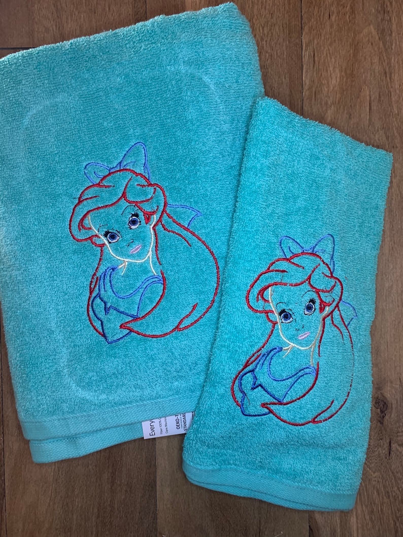 May include: Two turquoise terry cloth towels, one larger than the other, each featuring an embroidered outline of a long-haired character with a bow. The embroidery uses red, blue, and white thread to define the hair, facial features, and clothing.