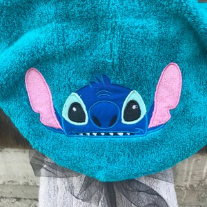 Op de afbeelding: Een fel turquoise handdoek met capuchon voorzien van een geborduurde applicatie van het personage Stitch. Het figuur heeft donkerblauwe vacht, grote roze binnenoren en lichtblauwe oogvlekken. Een laag zwarte tule is aan de onderkant bevestigd.