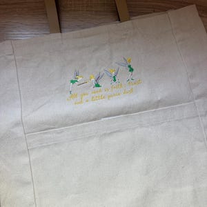 May include: A beige canvas tote bag with two brown handles. The front features an embroidered design of four small fairies in green dresses with wings, accompanied by the yellow embroidered text: All you need is faith, trust and a little pixie dust.