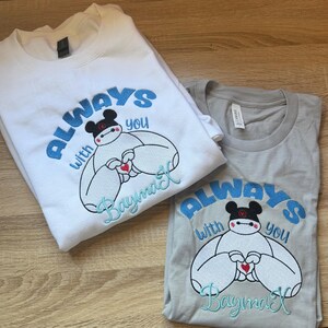 May include: A white sweatshirt and a gray t-shirt, both featuring an embroidered graphic of the character Baymax wearing black ears. The design includes the blue text "ALWAYS WITH YOU" and the name "Baymax" in light blue script with a red heart.