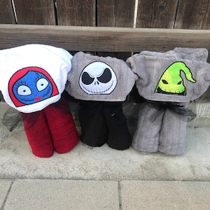K&ouml;nnte beinhalten: Drei Kapuzenhandt&uuml;cher mit gestickten Charaktergesichtern aus Nightmare Before Christmas. Das erste ist wei&szlig; mit einem blaugesichtigen Charakter, das zweite ist grau mit einem wei&szlig;en Totenkopfgesicht und das dritte ist grau mit einer gr&uuml;nen Geisterfigur.