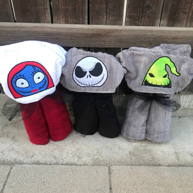 K&ouml;nnte beinhalten: Drei Kapuzenhandt&uuml;cher mit gestickten Charaktergesichtern aus Nightmare Before Christmas. Das erste ist wei&szlig; mit einem blaugesichtigen Charakter, das zweite ist grau mit einem wei&szlig;en Totenkopfgesicht und das dritte ist grau mit einer gr&uuml;nen Geisterfigur.