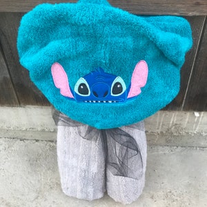 Op de afbeelding: Een fel turquoise handdoek met capuchon voorzien van een geborduurde afbeelding van het personage Stitch met grote roze oren en donkerblauwe gelaatstrekken. De handdoek is versierd met een zwart tule lint rond de basis van de capuchon.
