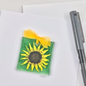 May include: A small, square, green glass ornament with a yellow sunflower painted on it. The ornament has a yellow ribbon tied around it.