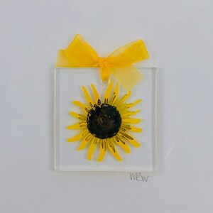 May include: A white card with a yellow ribbon tied around a clear square frame with a yellow sunflower inside. The card is signed "MRW".