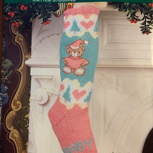 May include: Knitted baby stocking with a blue, white, and pink color scheme. The stocking features a small bear wearing a Santa hat and the word "BABY" is knitted into the stocking. The pattern includes hearts and Christmas trees. The stocking is 19 inches long.