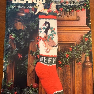 May include: A red and green knitted stocking with a Canada Goose design. The stocking is 19 inches long and has the name "JEFF" knitted on the front. The stocking is hanging on a wooden mantle with greenery and red berries.