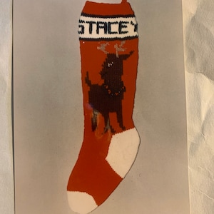 May include: A red and white knitted stocking with a black dog design and the name "STACEY" on the cuff. The stocking is part of a YarnCraft kit.
