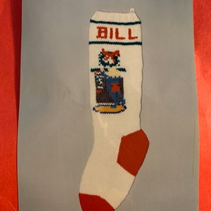 May include: White knitted Christmas stocking with red and blue accents. The stocking has the name "BILL" embroidered on the top and a cartoon image of a person in a blue and brown outfit.