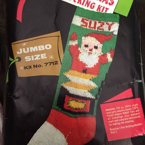 May include: A Christmas stocking knitting kit with a red and green stocking featuring a Santa Claus design. The kit includes 24 ounces of yarn, sequins, beads, French Angora Rabbit yarn, and all trimmings. The kit also includes an alphabet chart and instructions for making one stocking. The kit is labeled "Jumbo Size" and "Kit No. 7712".