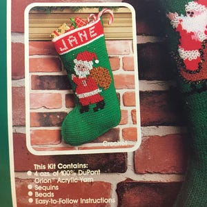 May include: A green and red crochet stocking with the name "JANE" embroidered on the front. The stocking features a Santa Claus figure holding a bag of gifts. The kit includes 4 ounces of 100% DuPont Orion Acrylic yarn, sequins, beads, and easy-to-follow instructions.