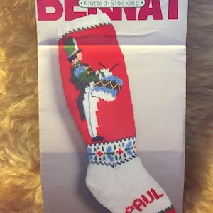 May include: A red and white knitted stocking with a drummer boy design. The stocking has a white cuff with blue and green accents and the name "PAUL" embroidered on the toe. The stocking is labeled "Bernat Knitted Stocking" and "W00622 DRUMMER".