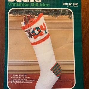 May include: A Bucilla Christmas stocking kit to knit, featuring a white stocking with red and green stripes, a red bow, and the word "Joy" in green letters. The kit includes 21 1/2 ounces of yarn, 12 inches of red velvet ribbon, and easy-to-follow instructions. The stocking is 20 inches high.