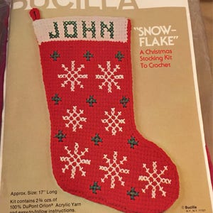 May include: A red and white crochet stocking kit with a snowflake design. The kit includes yarn and instructions to crochet a stocking that is 17 inches long. The stocking is personalized with the name "John".
